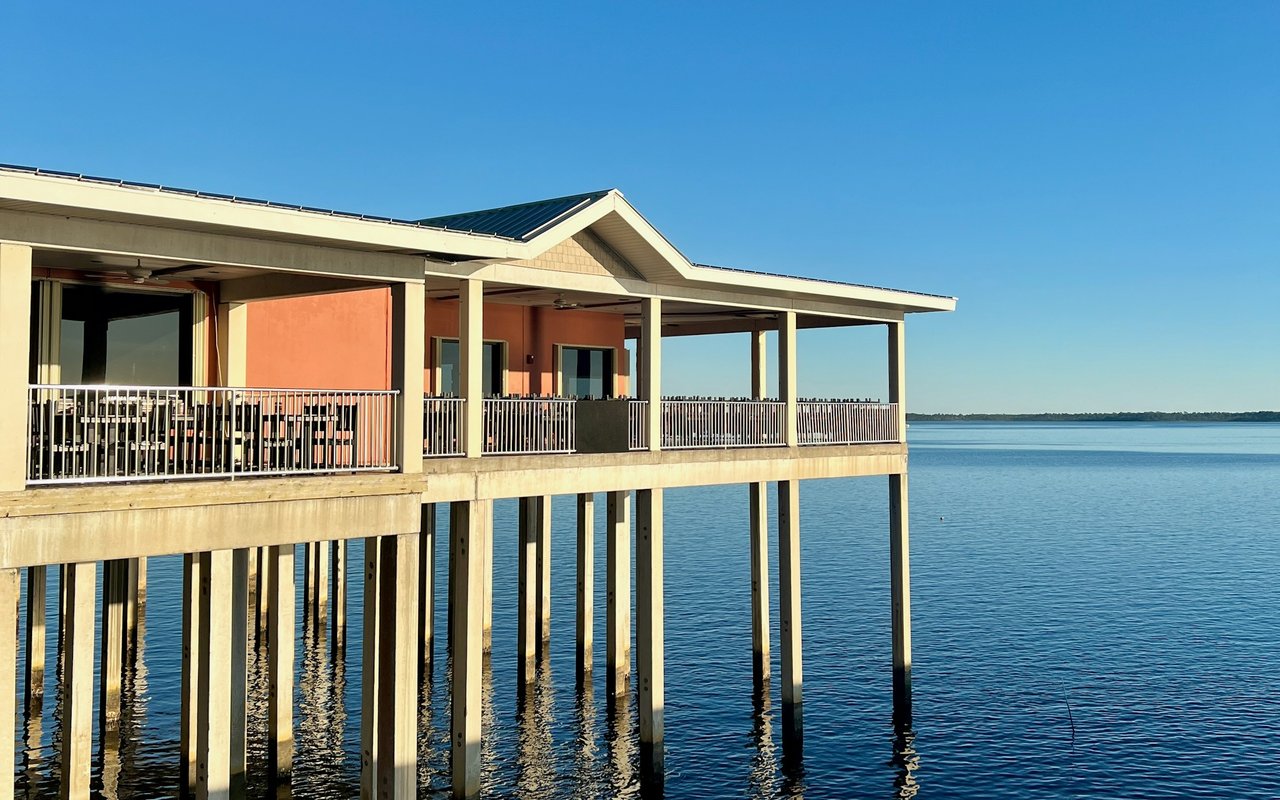 Waterfront building raised on pilings, with a long covered deck over calm water.