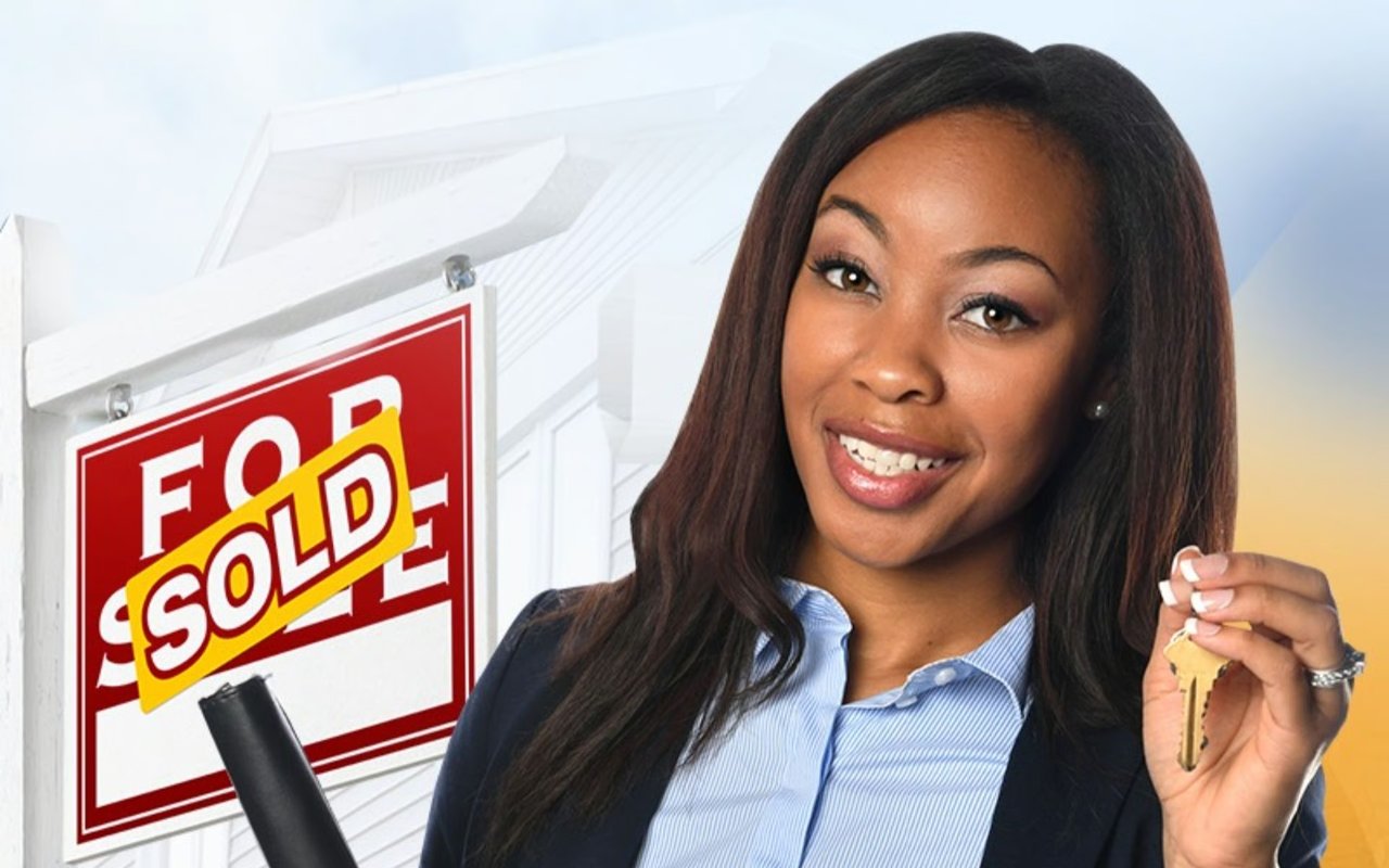 Smiling woman holds house keys beside a real estate sign marked “SOLD.”