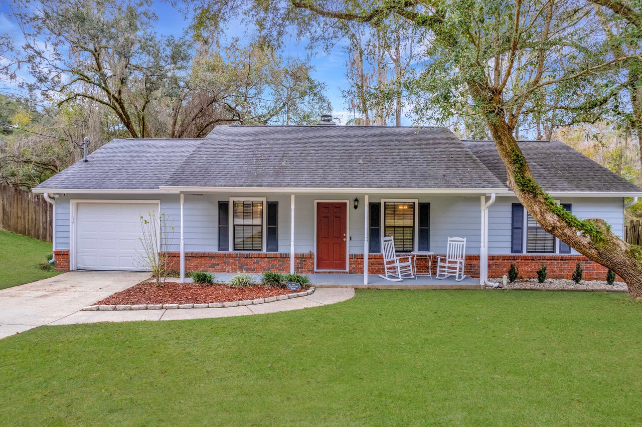 Single-story house with blue siding, brick accents, and a red door. Front porch has white rocking chairs. Surrounded by green lawn and trees.