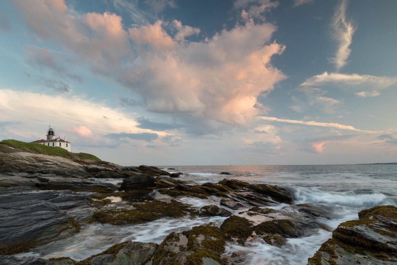 Lighthouse above a rocky Jamestown shoreline, with waves and pink clouds at sunset.