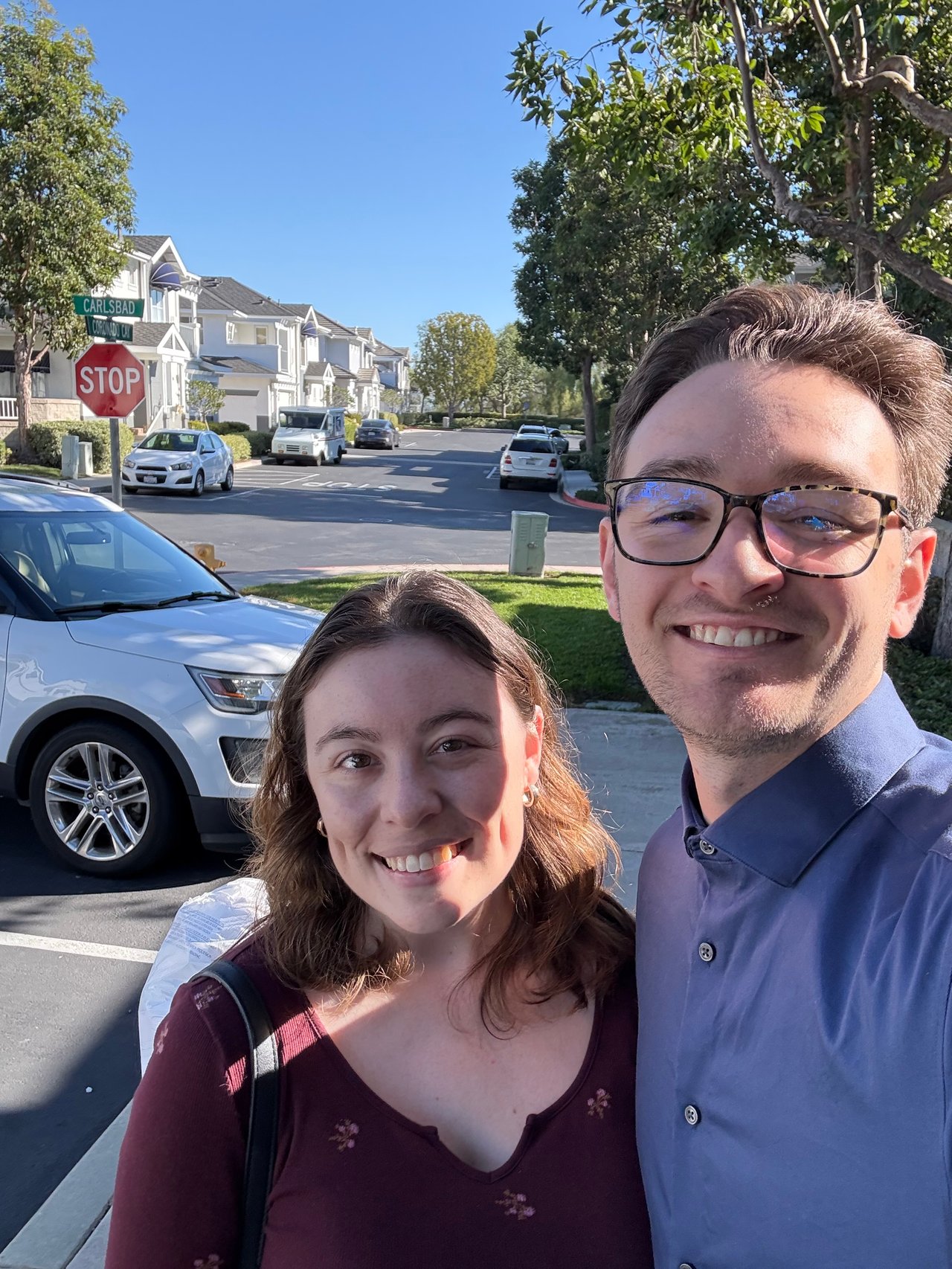 Two smiling adults pose for a selfie on a sunny residential street.
