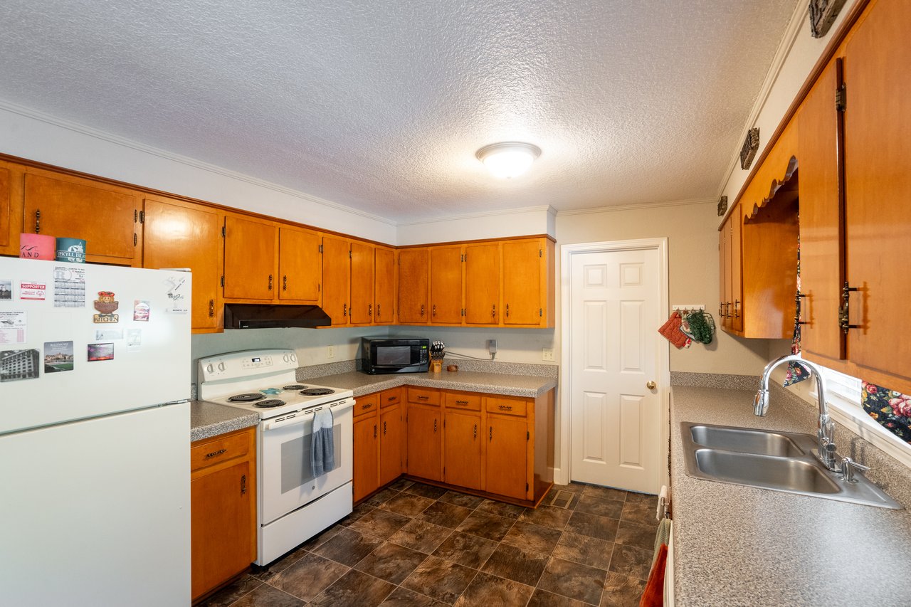 A charming kitchen with wooden cabinets, a white refrigerator, stove, and microwave on a countertop. There's a double sink and a door on the right. Perfect for real estate agents showcasing cozy homes, it boasts dark tile flooring ideal for prospective buyers.