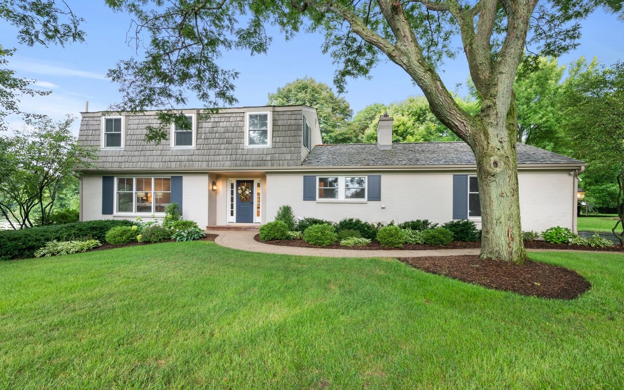 White house with a shingle-clad upper level, blue shutters, landscaped garden, and large shade tree.