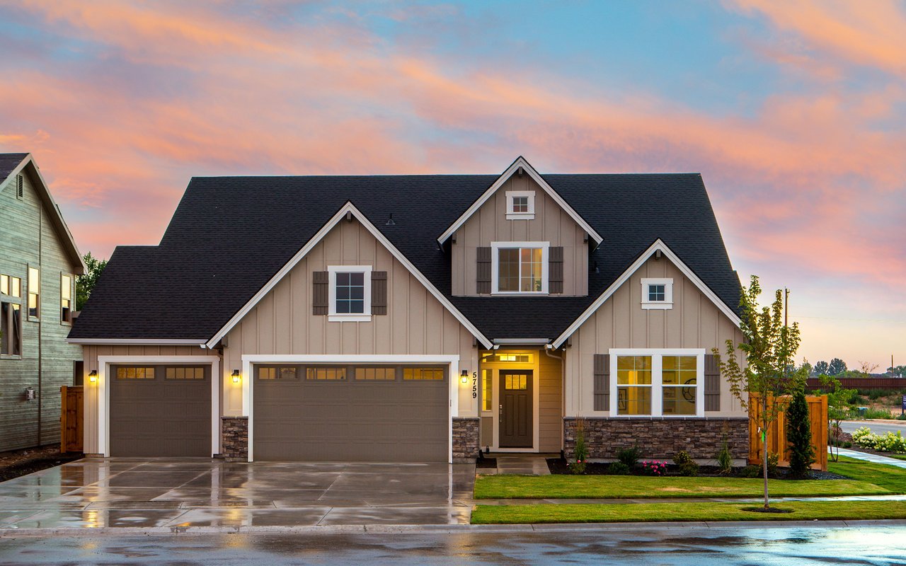 Taupe house with two garage doors and a landscaped lawn beneath a pink sunset sky.
