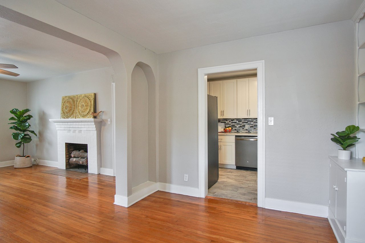 Spacious living room with hardwood floors and a white brick fireplace. Arched entry leads to a modern kitchen with white cabinets and a mosaic backsplash.