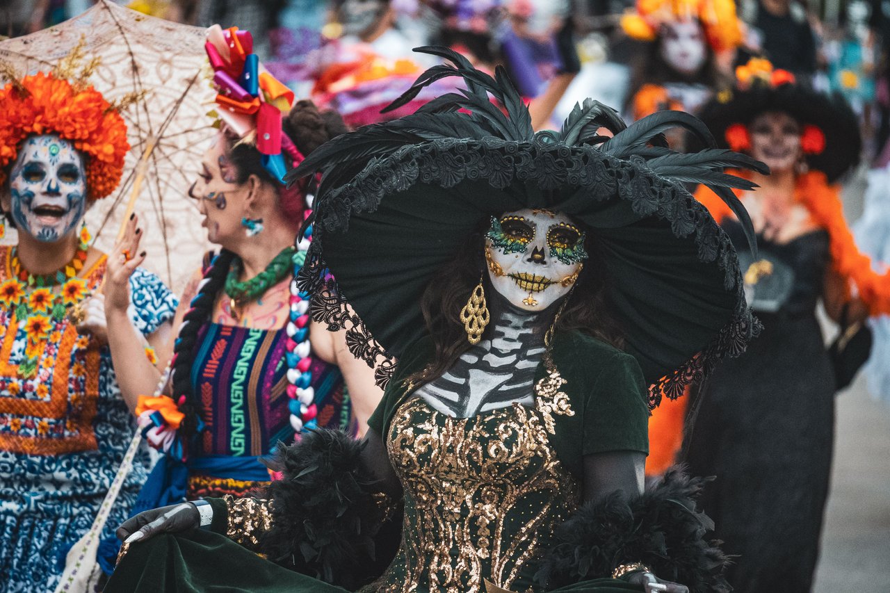 A wide photograph of the Village Halloween Parade on Sixth Avenue, with costumed participants and Manhattan buildings in the background.