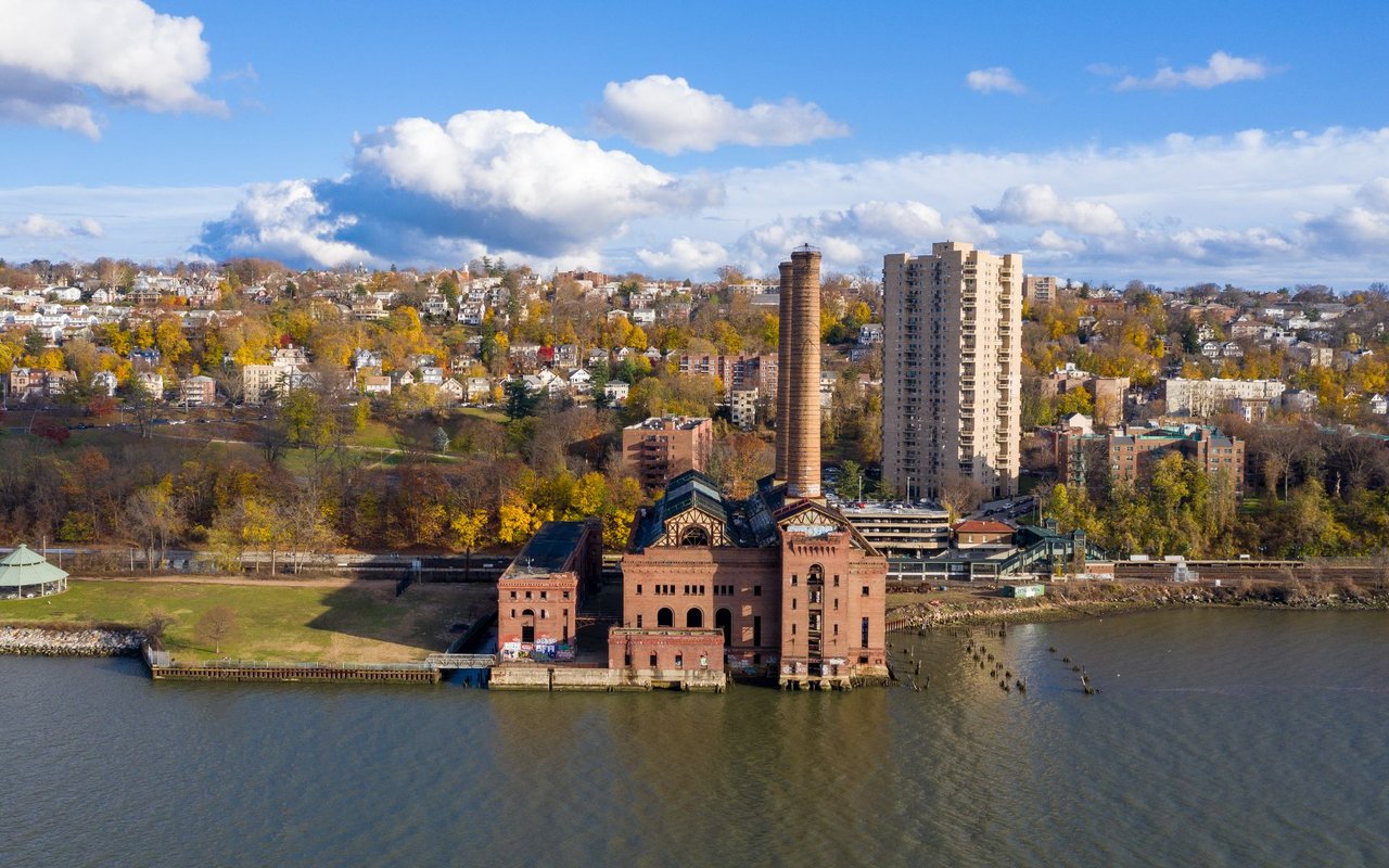 Yonkers waterfront with a brick industrial building, apartment towers, and autumn foliage along the hillside.