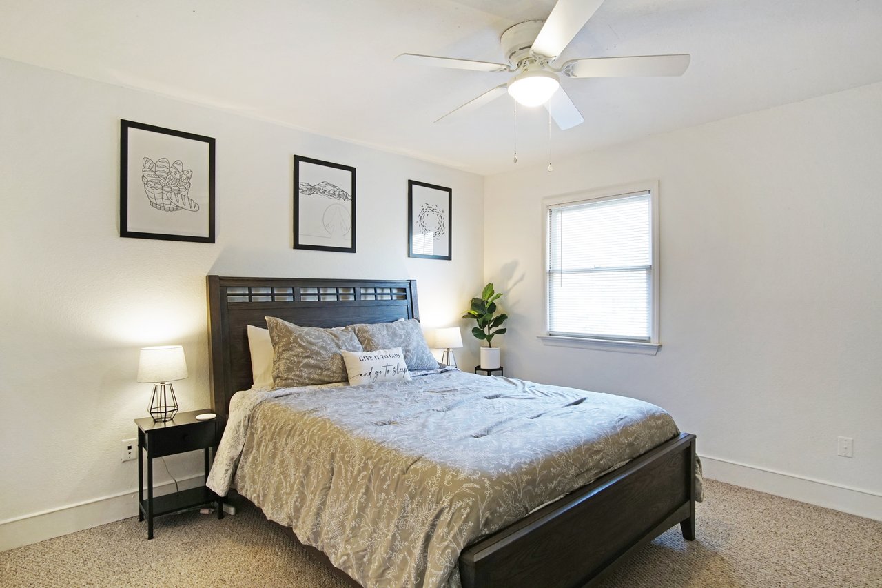 A serene bedroom with a gray-patterned bedspread, modern lamps, and minimalistic wall art. A ceiling fan and potted plant add a cozy touch.