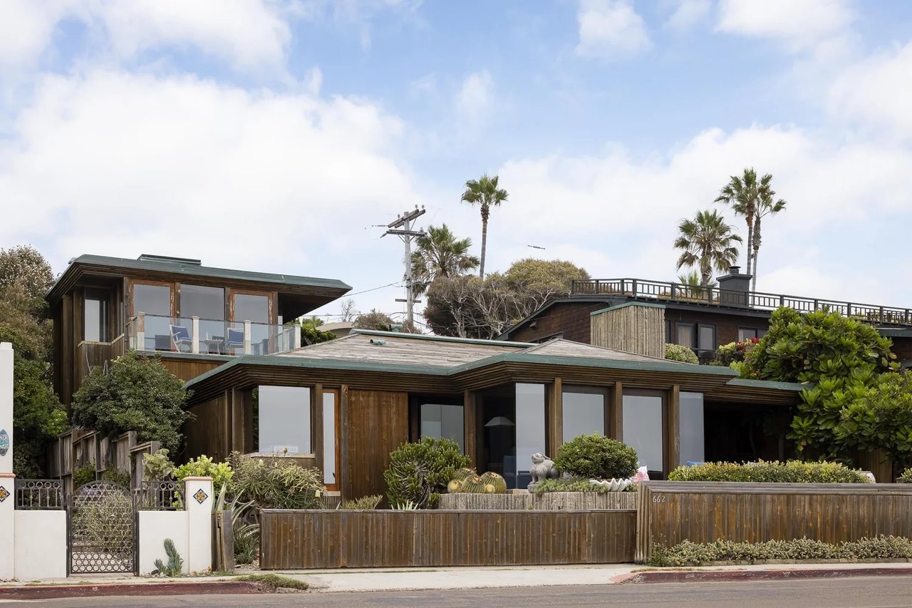 Wood-clad modern home with broad windows, an upper balcony, and a fenced, landscaped front yard.