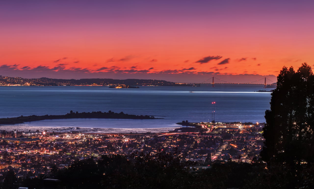 City lights and a suspension bridge across the bay beneath a vivid sunset sky.
