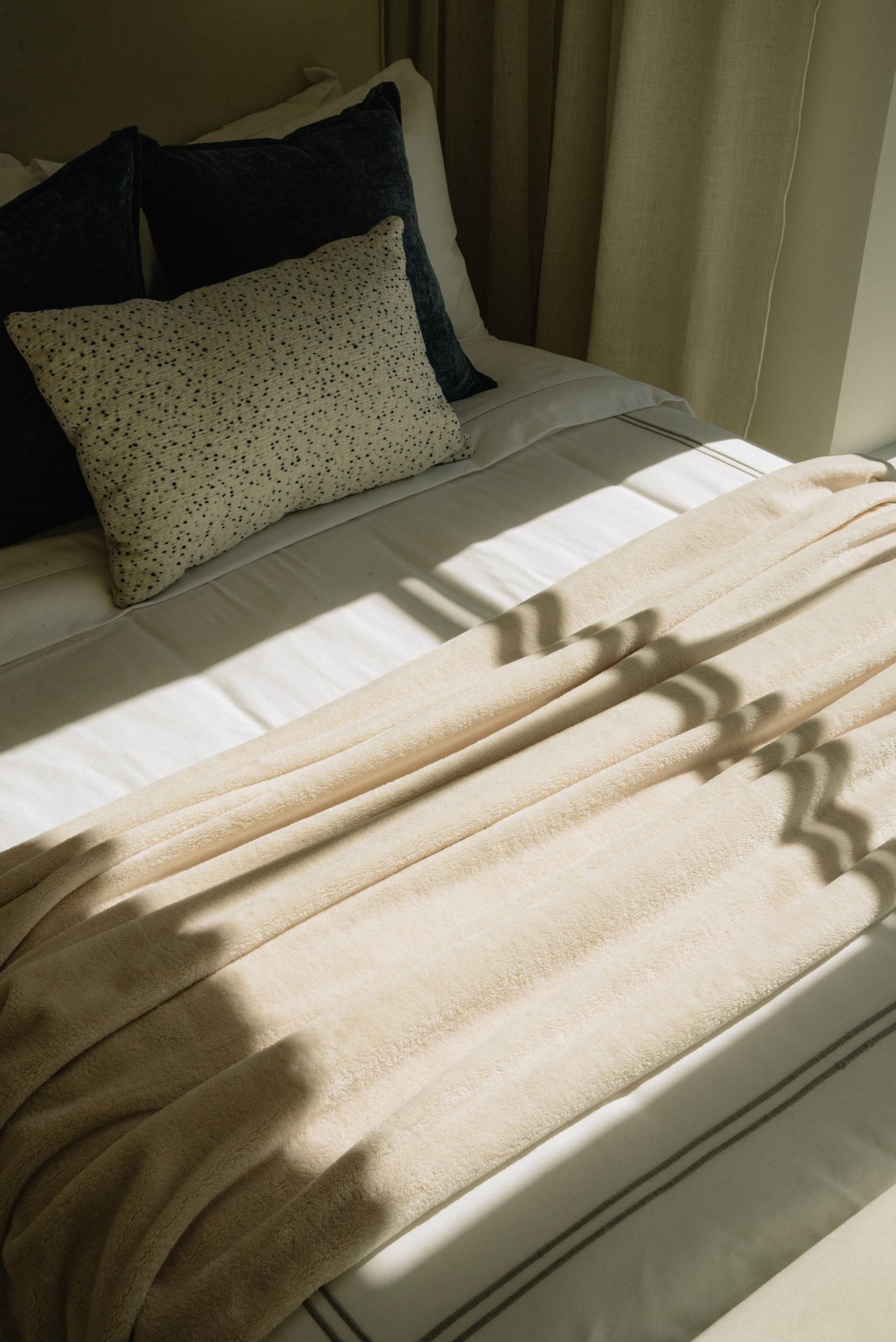 Staged bedroom with a plush cream blanket and patterned pillows in sunlight.