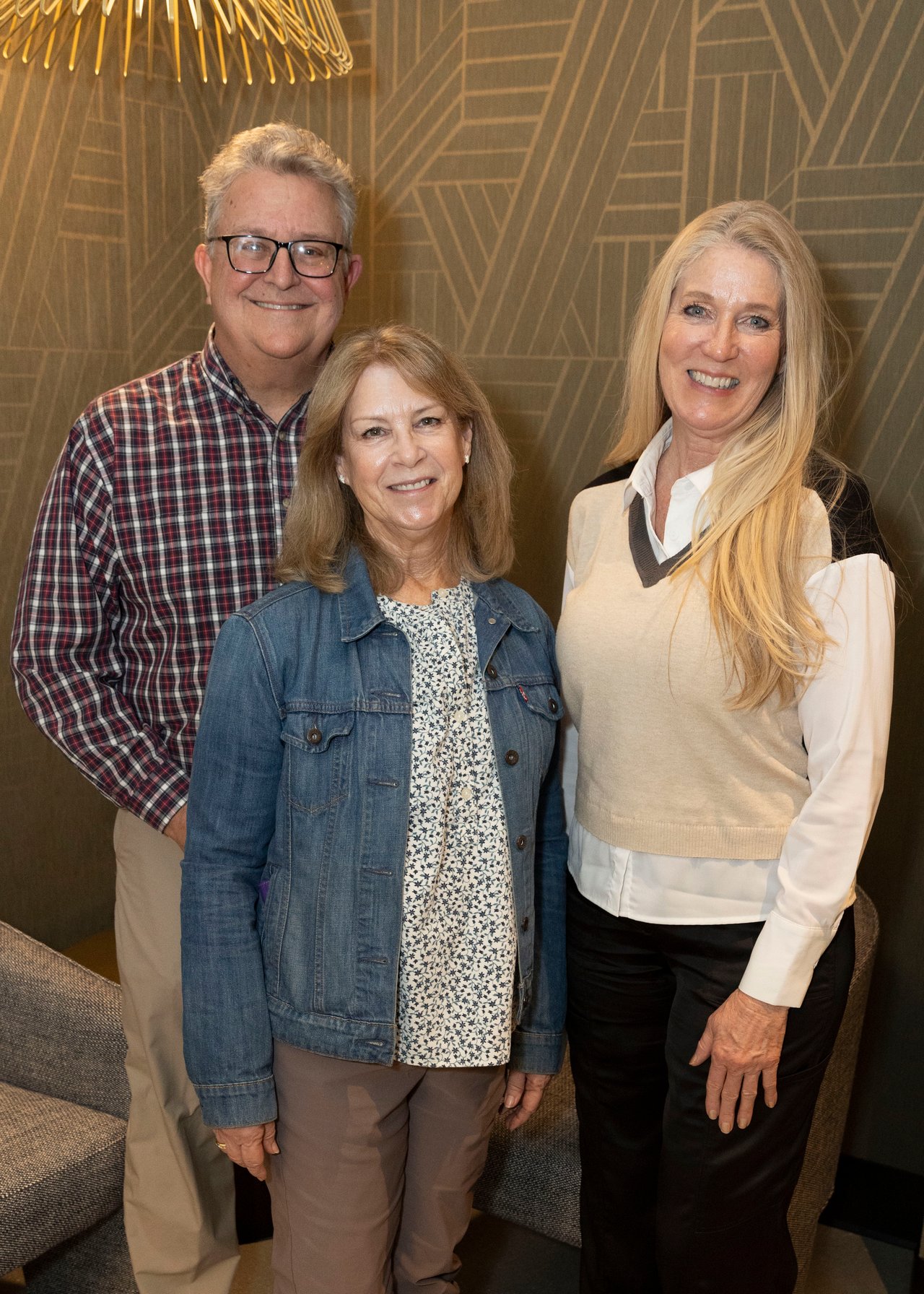 Three smiling adults pose together indoors, including a man in glasses and two women.