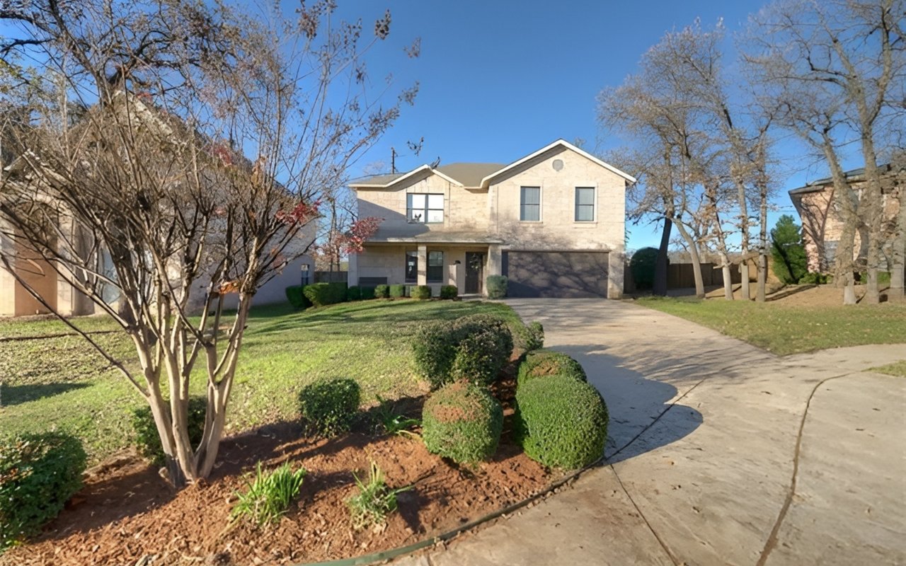 Two-story stone house with a double garage, curved driveway, and neatly trimmed shrubs.