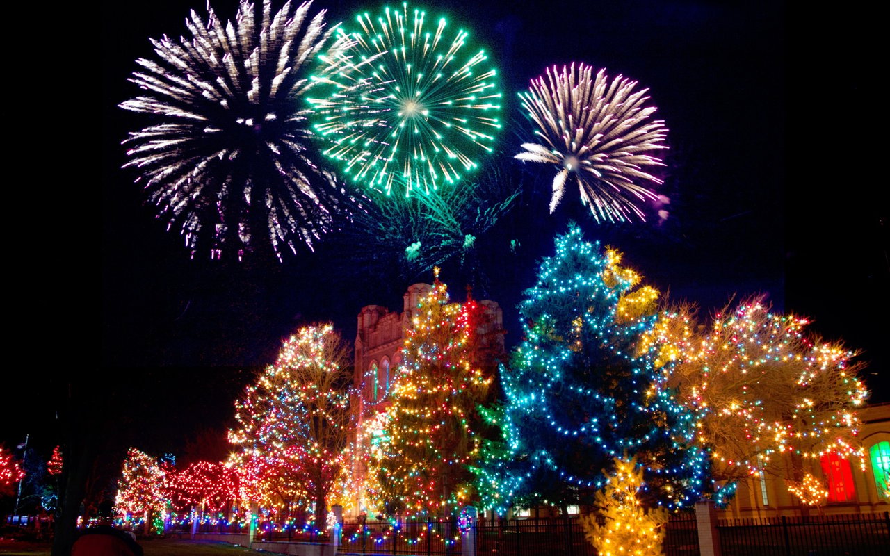 Colorfully lit Christmas trees beneath fireworks at a nighttime holiday festival.