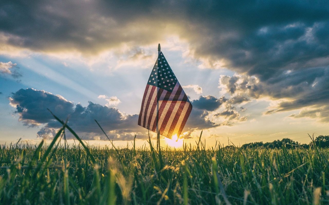 American flag in a grassy field, silhouetted against a dramatic sunset sky.