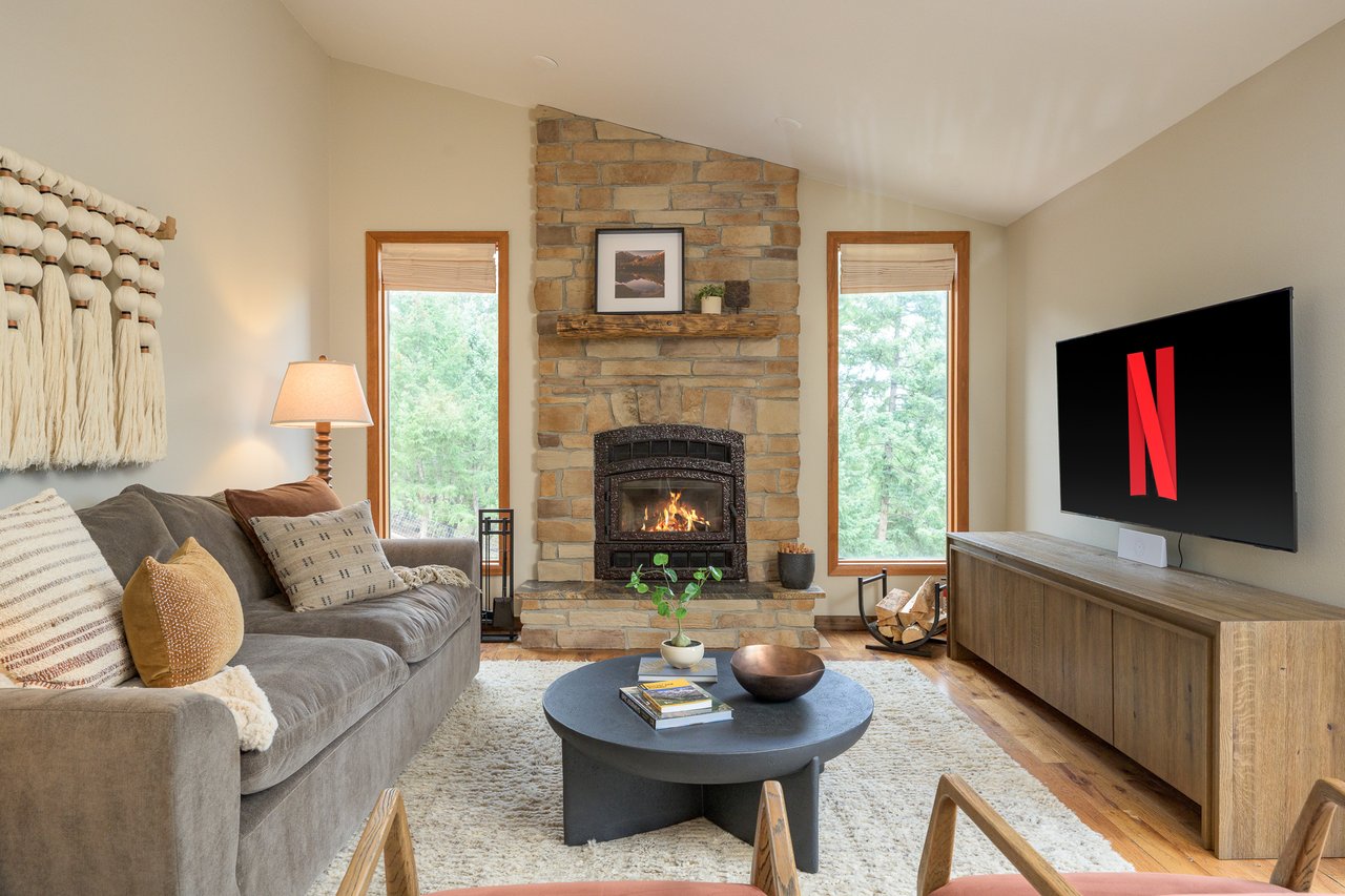 Living room with a tall stone fireplace, vaulted ceiling, and windows overlooking trees.