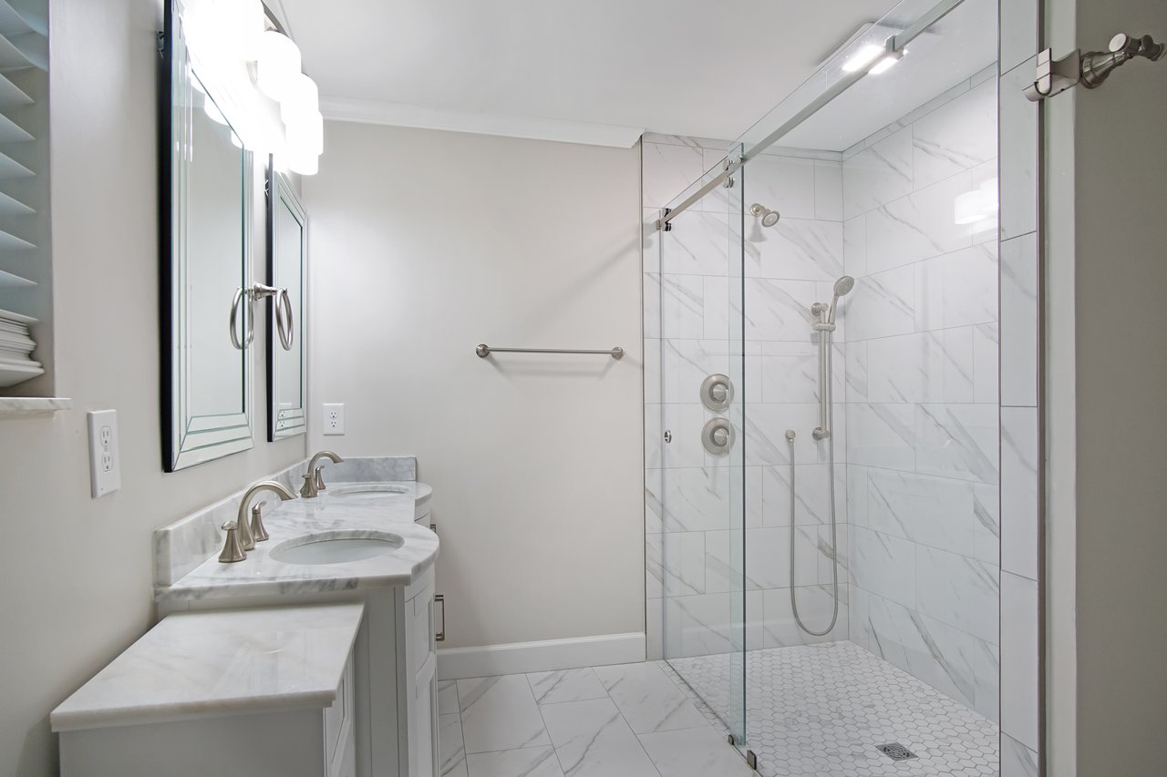 Elegant bathroom with marble tiles, a double sink vanity with curved edges, and a glass-enclosed shower. Minimalist and serene atmosphere.