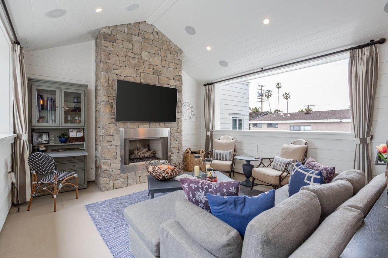  A modern sunroom with a floor-to-ceiling stone fireplace, a large TV, a gray sectional sofa, and a wide window.
