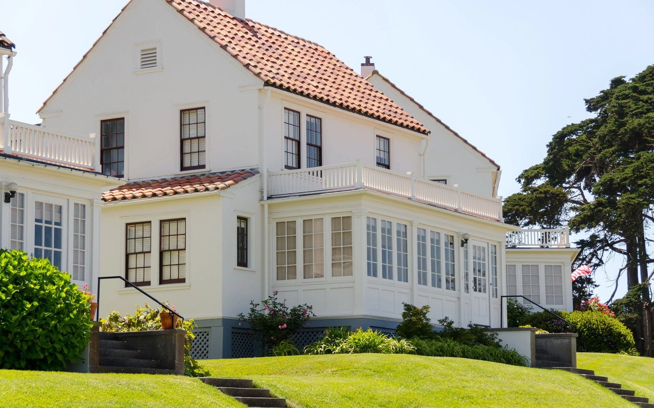 White multi-story house with a red tile roof, balconies, and a landscaped lawn.