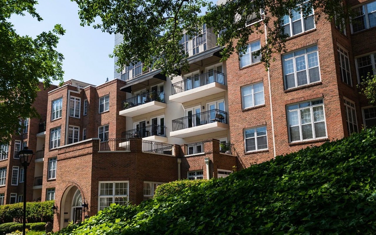 Brick apartment building with balconies, leafy trees, and landscaped hedges.