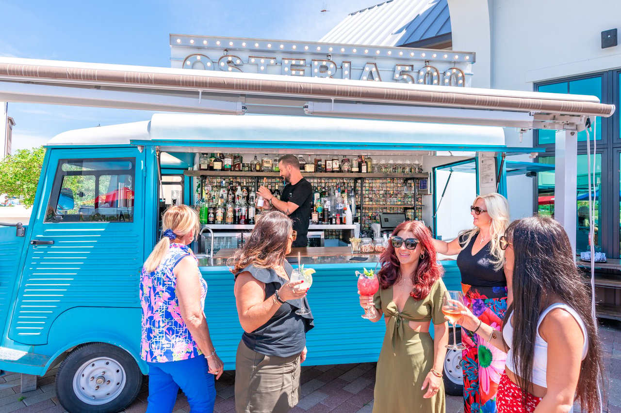 Women socializing with cocktails beside a turquoise food truck at an outdoor shopping destination.