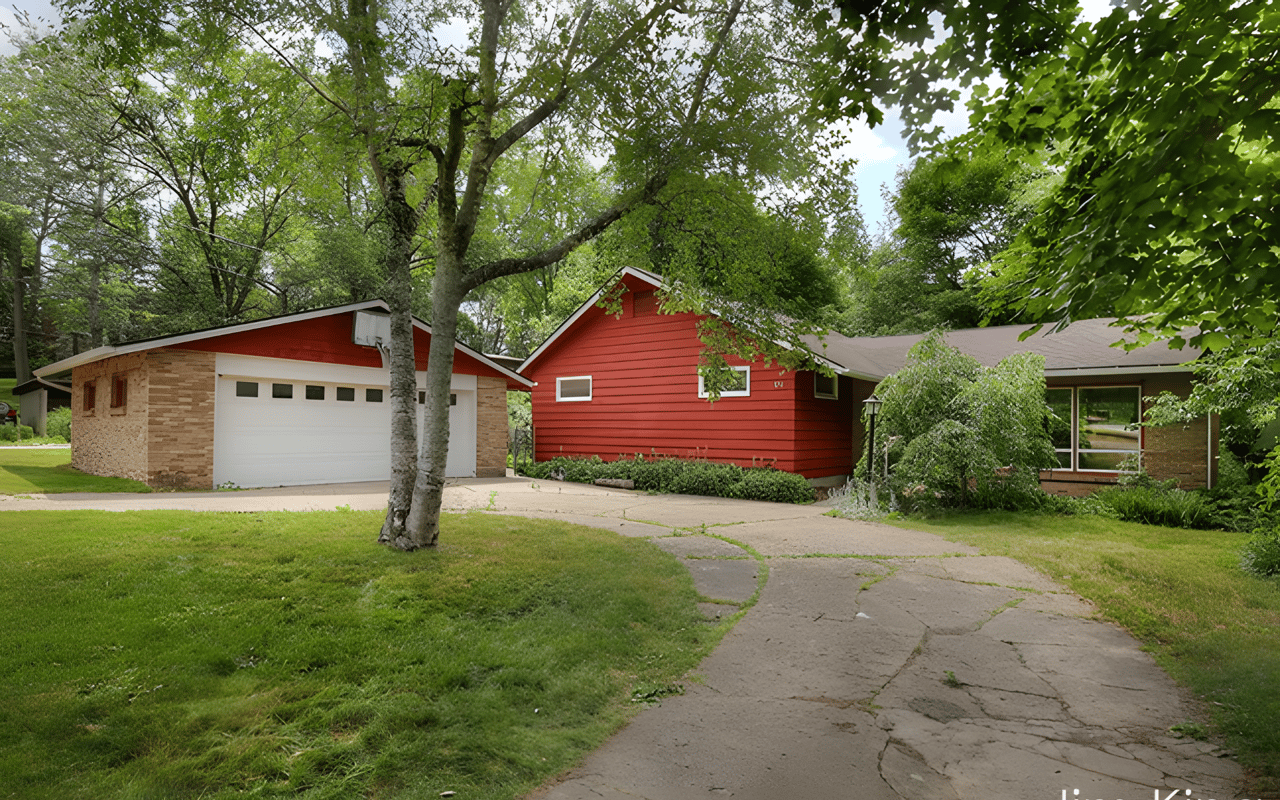 Single-story red-sided home with an attached garage, mature trees, and a paved driveway.