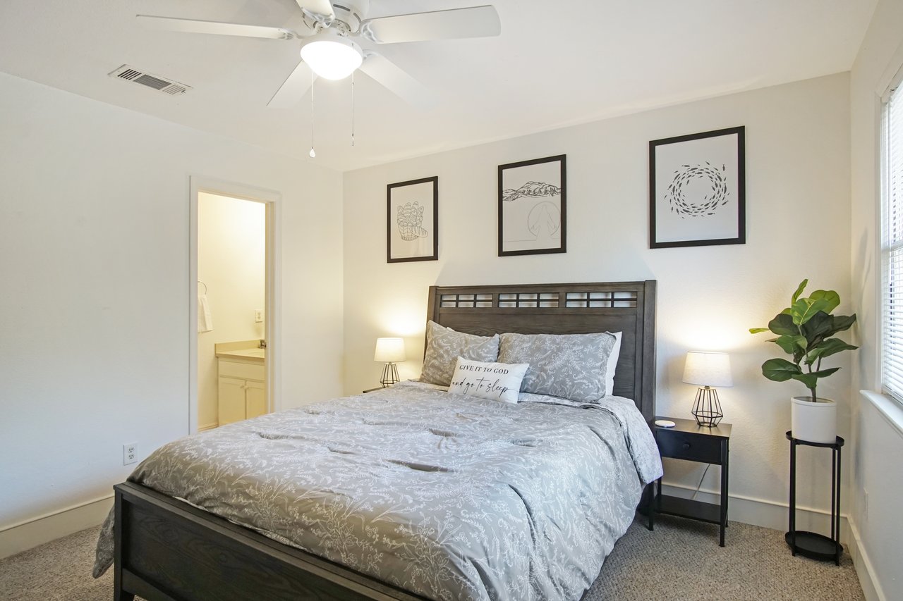 Bedroom with a gray-patterned bed, three framed artworks, and a potted plant by the window. Warm lighting and a ceiling fan create a cozy atmosphere.Bedroom with a gray-patterned bed, three framed artworks, and a potted plant by the window. Warm lighting and a ceiling fan create a cozy atmosphere.