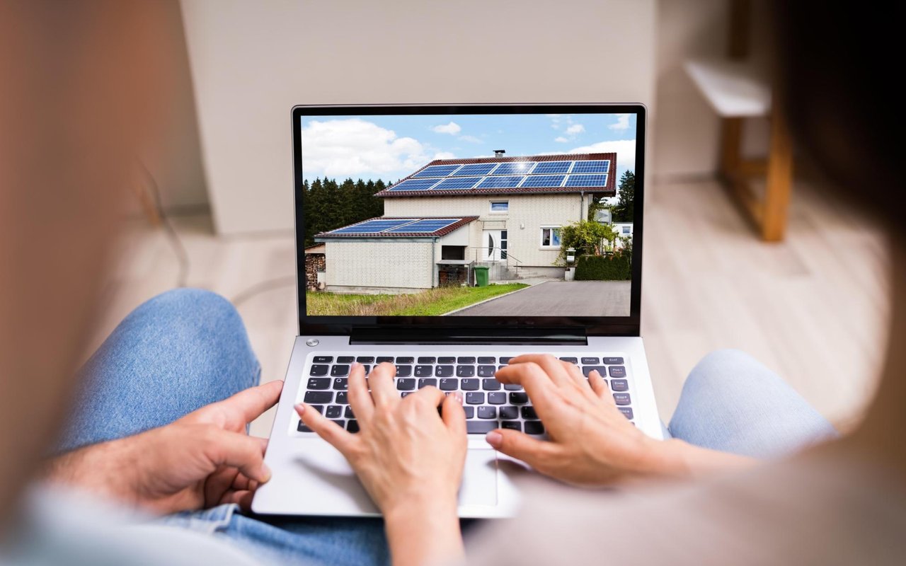 A person views a solar-panelled house on a laptop while seated at home.
