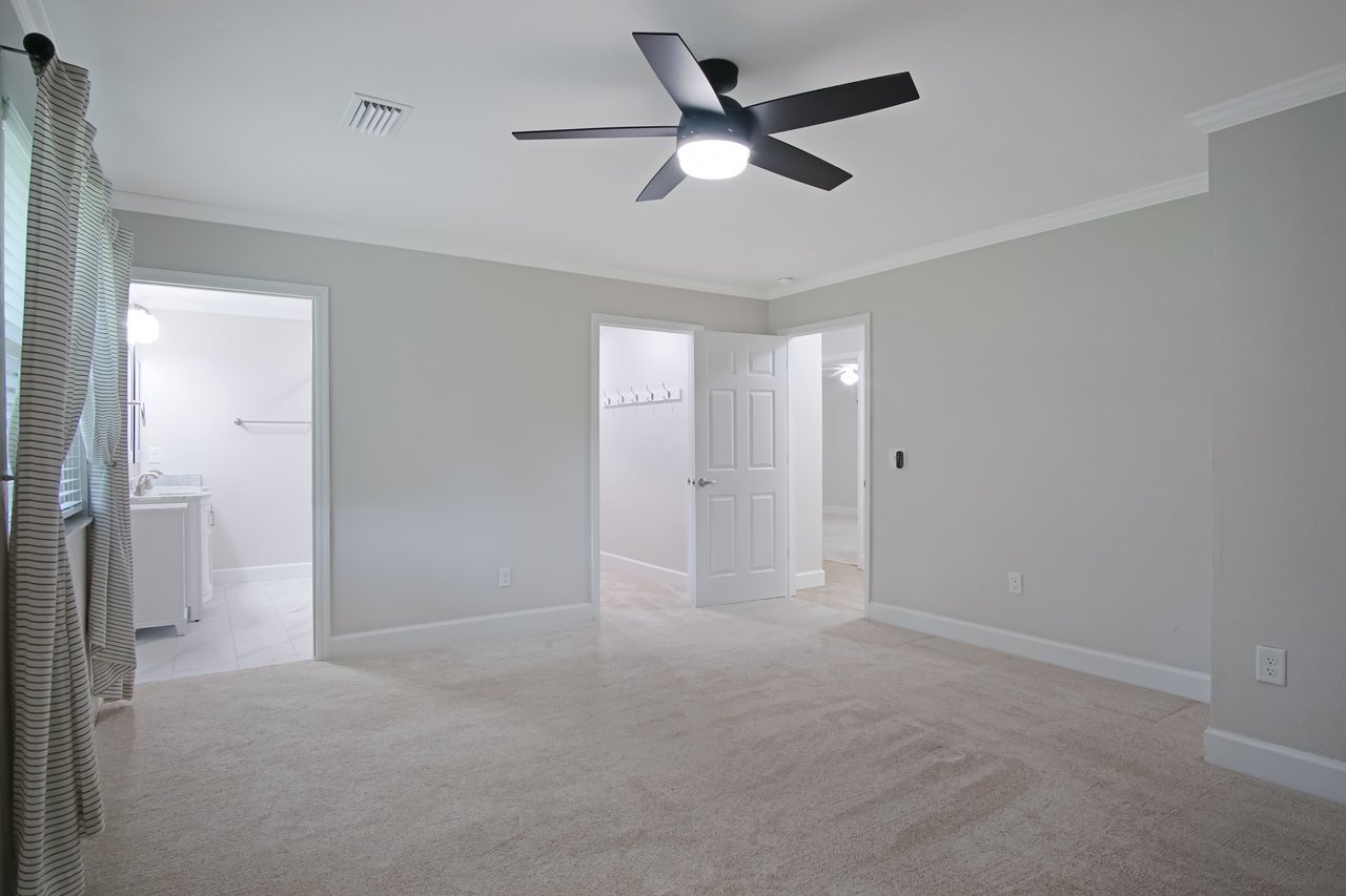 Empty room with beige carpet and light gray walls, featuring a black ceiling fan, striped curtains, and white doors leading to adjacent rooms.
