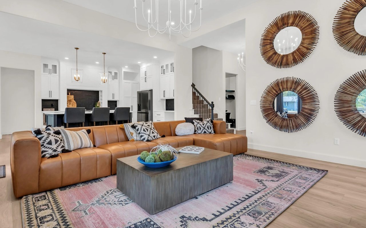 Open-plan living room with tan sectional, pink patterned rug, sunburst mirrors, and white kitchen.