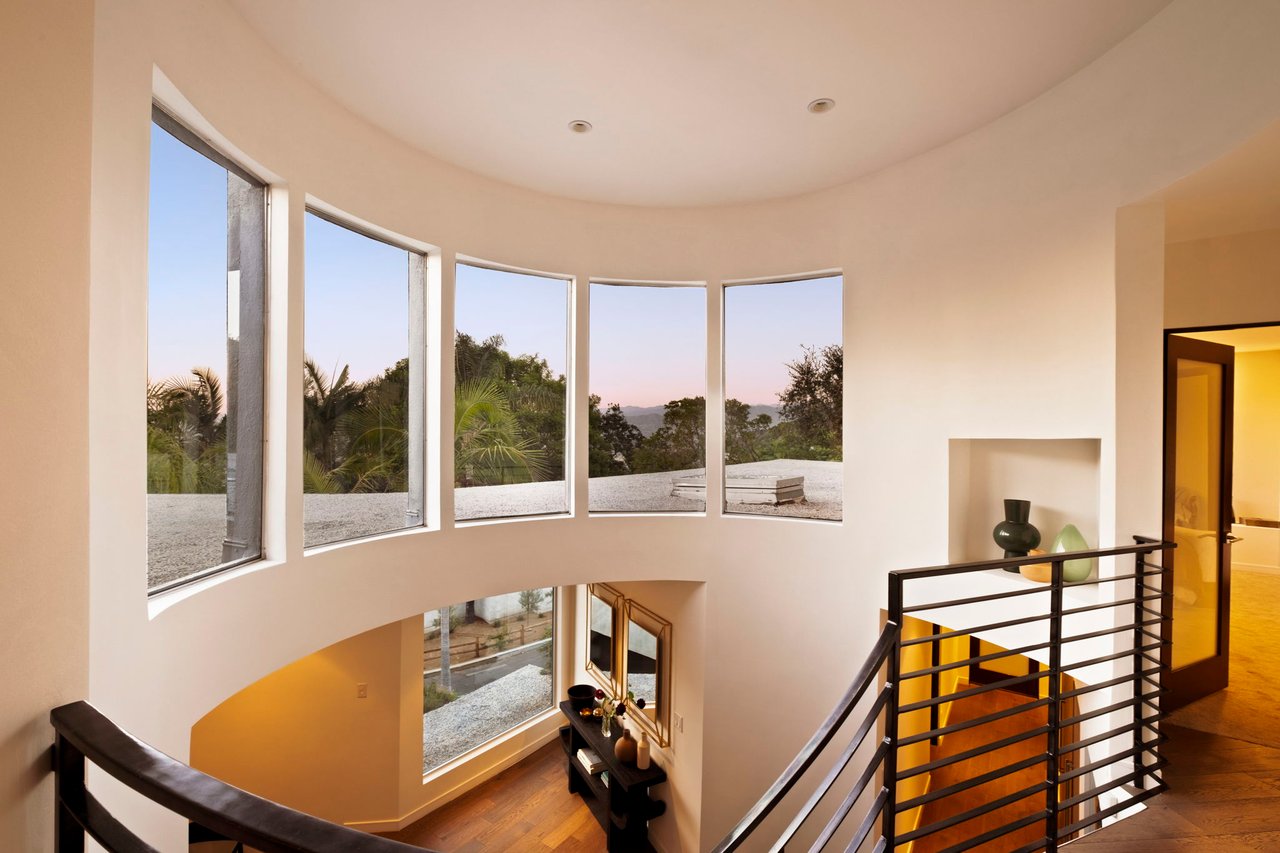 Double-height entry with curved picture windows overlooking tree-covered hills and a black metal staircase railing.