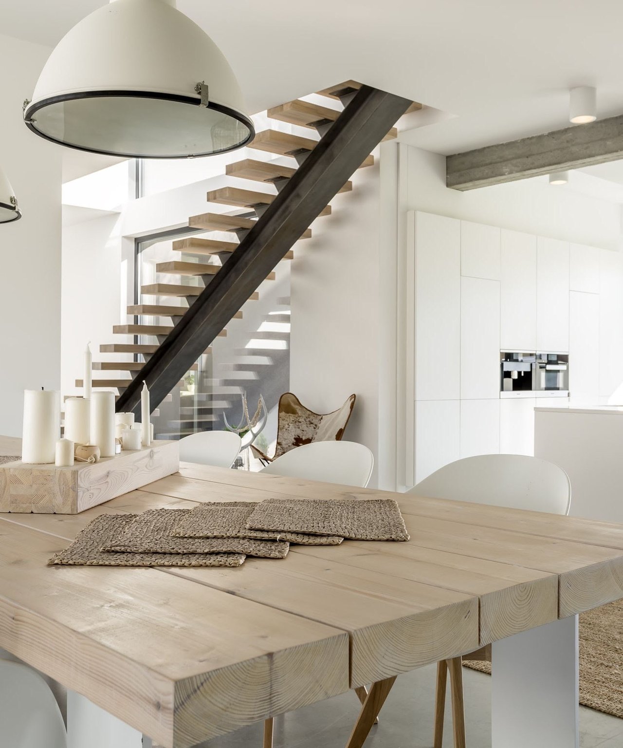 Modern dining area with a pale wood table and an open-riser staircase beside a white kitchen.