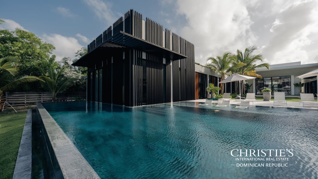 Modern villa with a black slatted pavilion beside a large turquoise pool and palm trees.