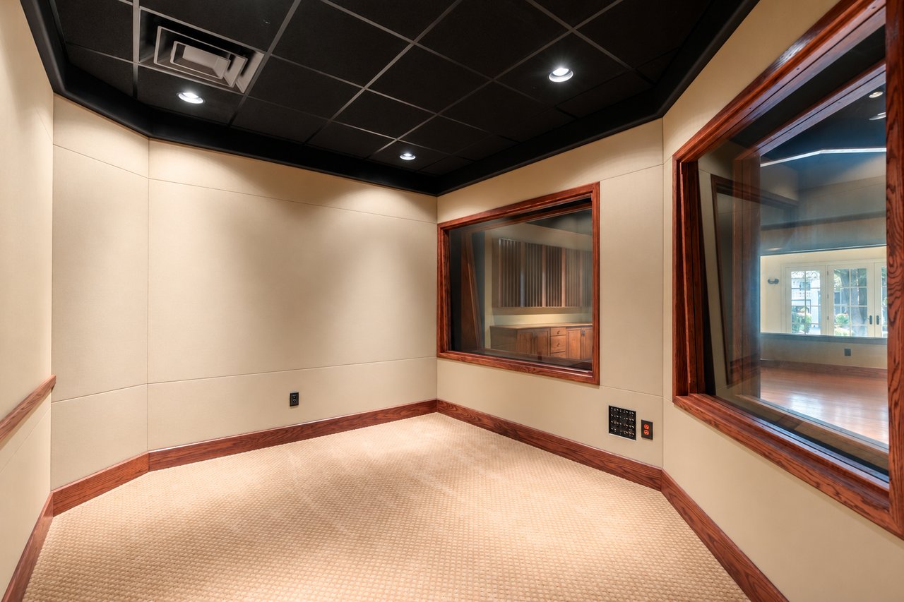 Sound-treated recording room with beige acoustic panels and wood-trimmed windows into adjoining studio spaces.