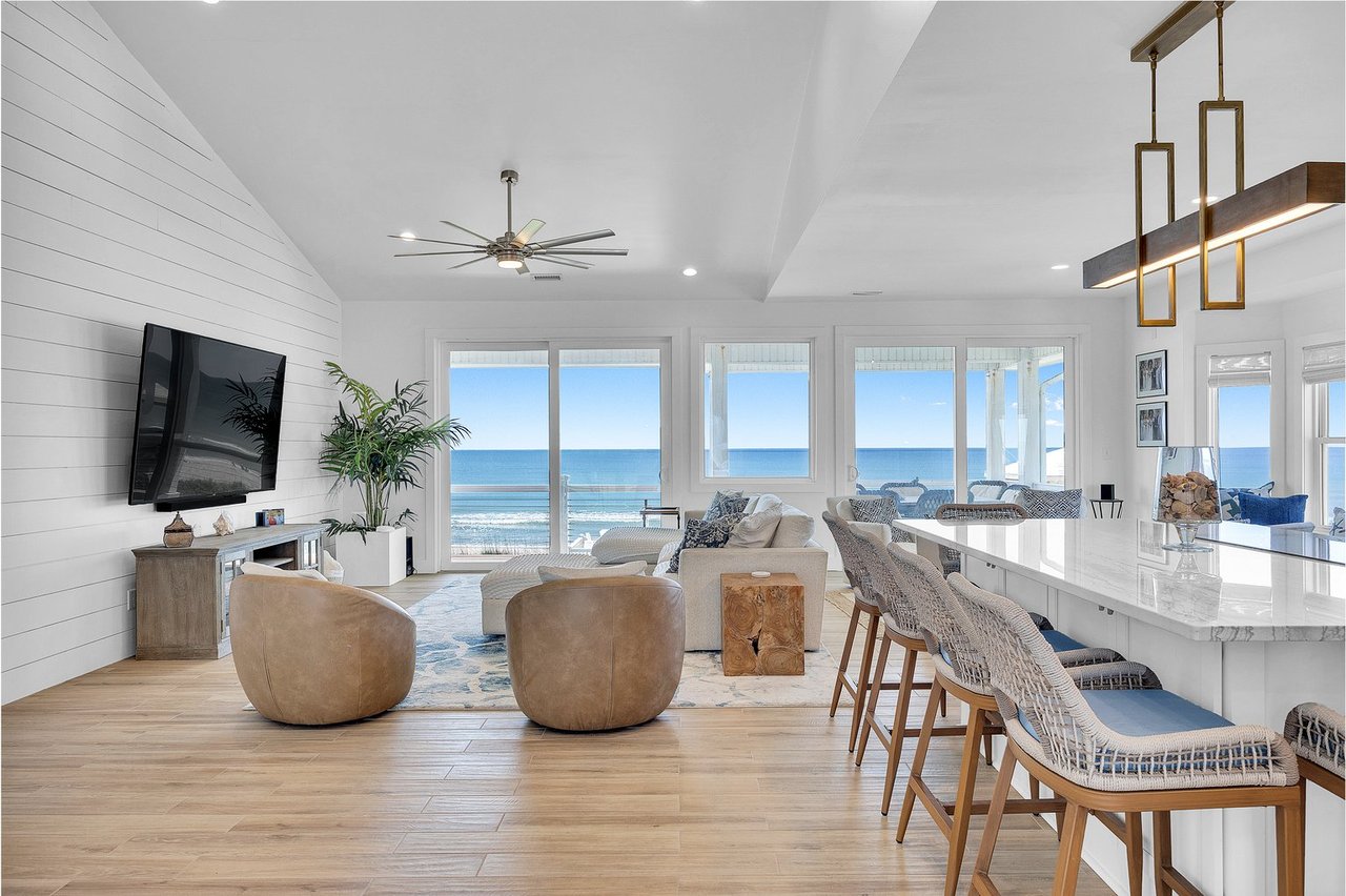Oceanfront living room with an open kitchen, woven bar stools, and wide windows overlooking the water.