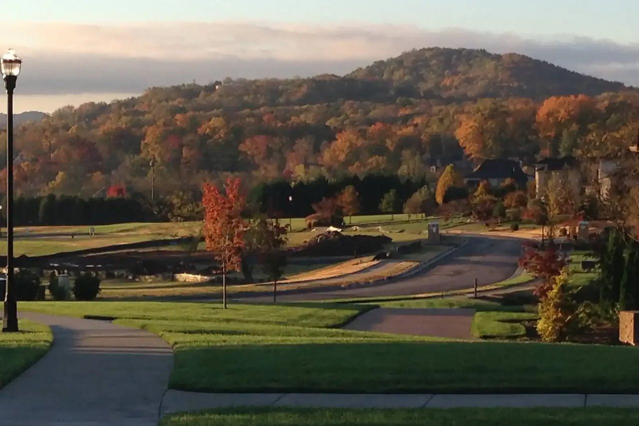 Autumn neighborhood with a curving road, landscaped lawns, and wooded hills beyond.