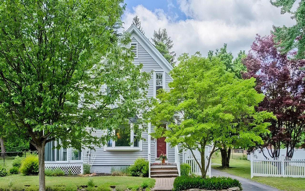 Light-gray gabled house with a red front door, leafy trees, and a white picket fence.