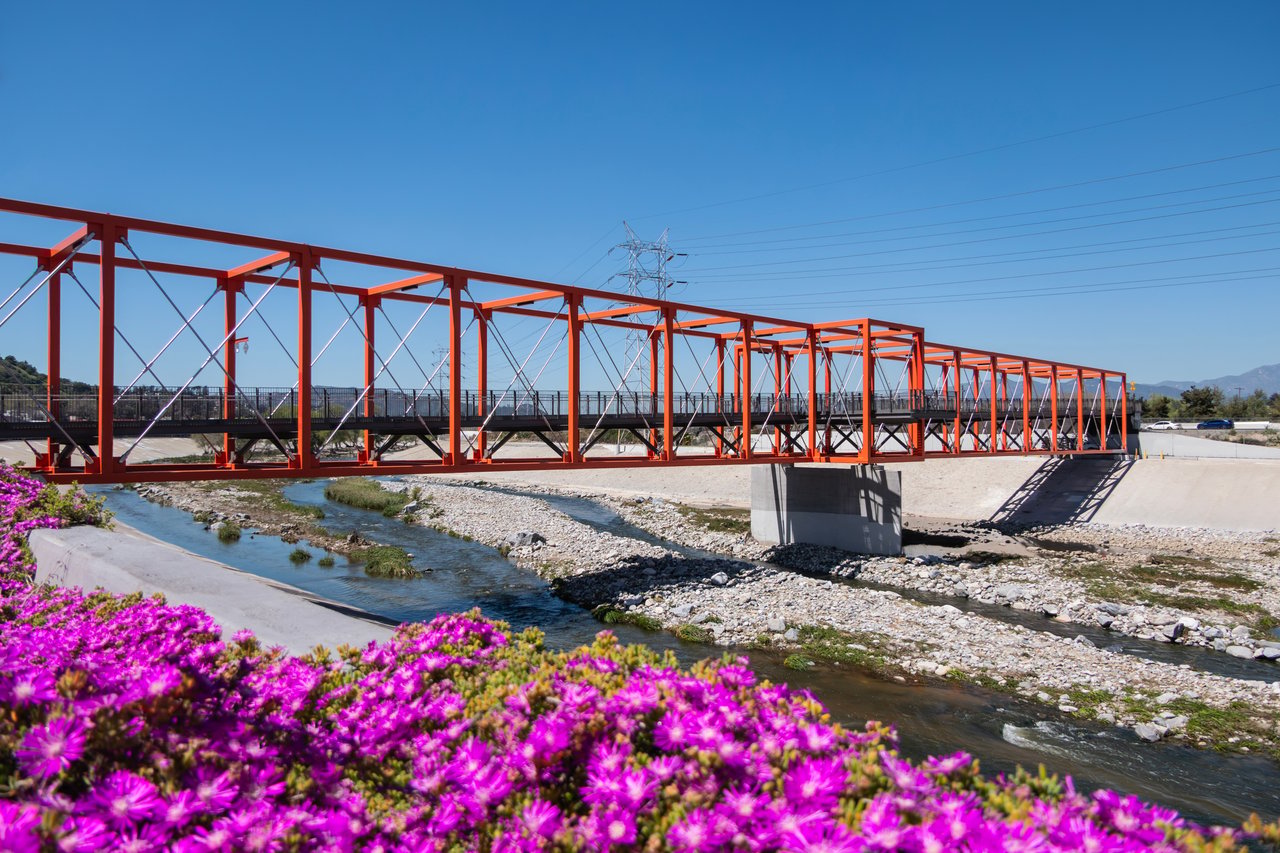 Red pedestrian bridge over the Los Angeles River in Elysian Valley, with magenta flowers in the foreground.
