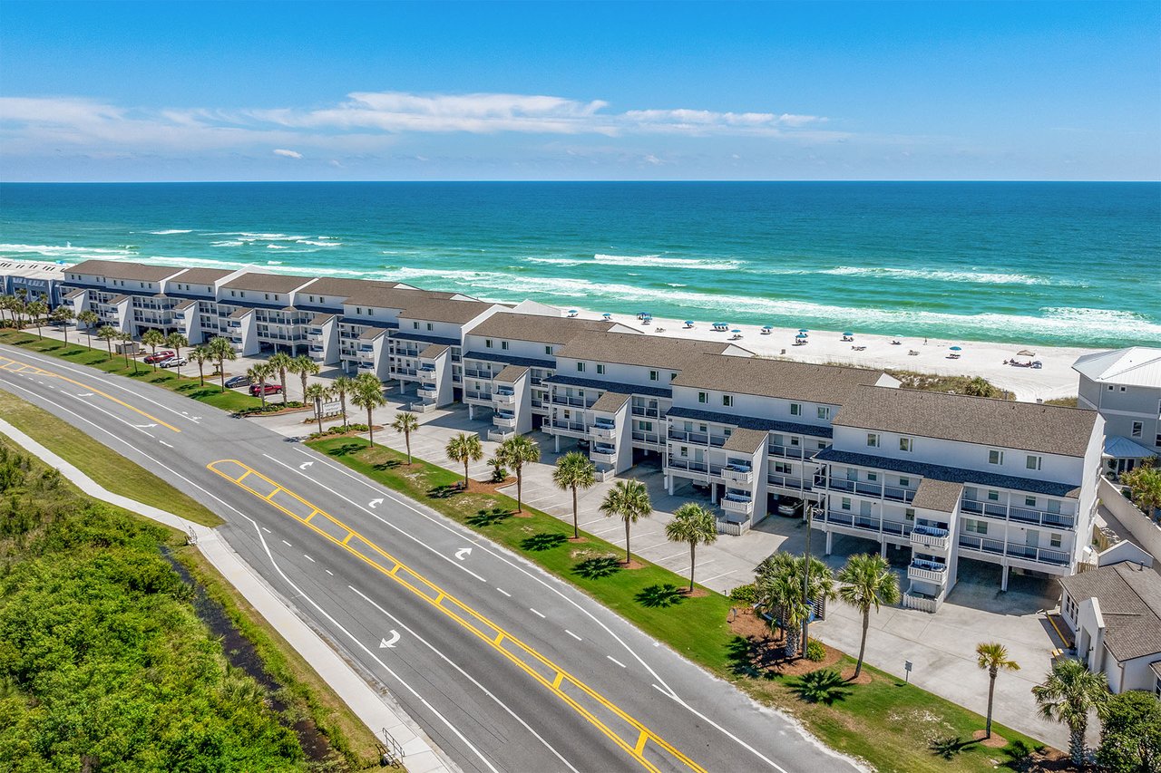 Aerial view of beachfront condominiums beside a coastal road, sandy beach, and Gulf waters.