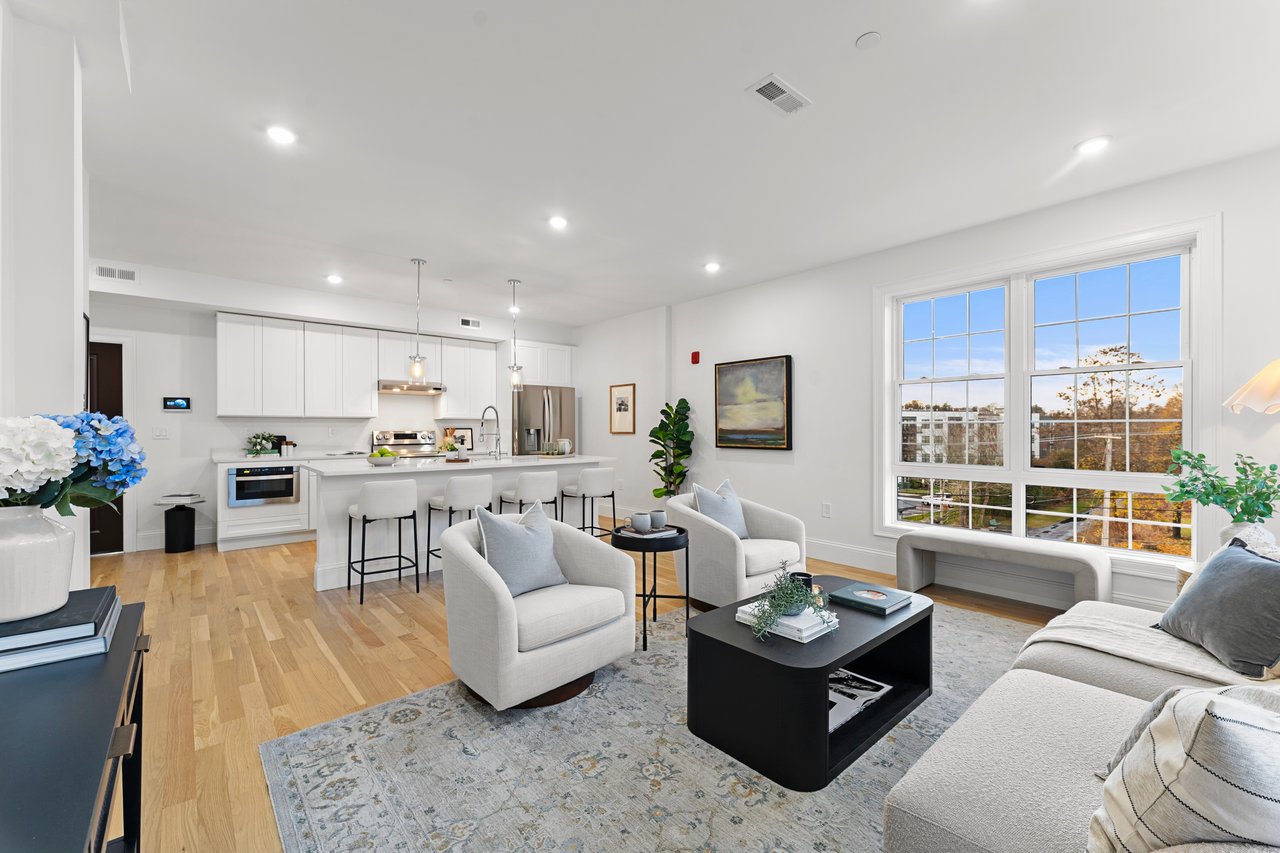 Open-plan apartment living room and kitchen with white cabinetry, light wood floors, and large windows overlooking autumn trees.