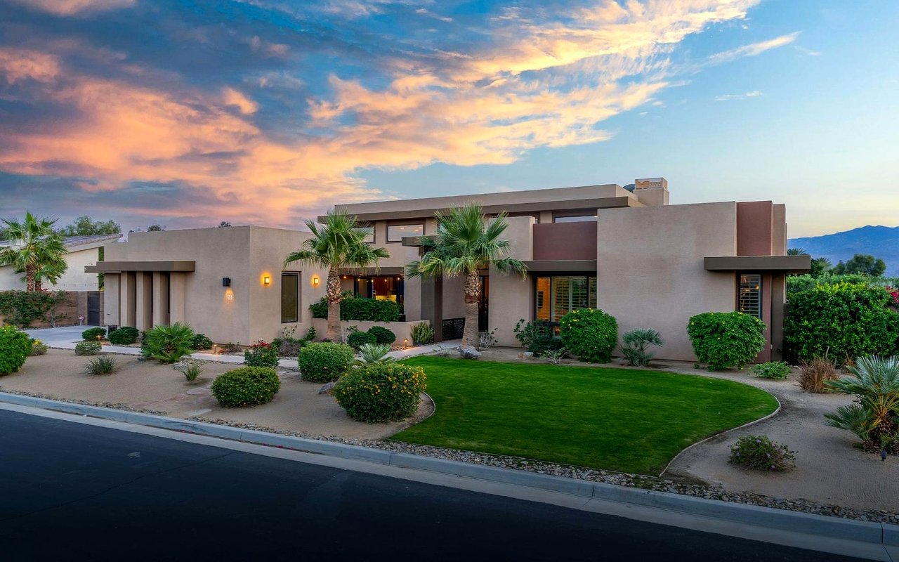 Modern stucco home with palm trees, curved lawn, distant mountains, and a colorful sunset sky.