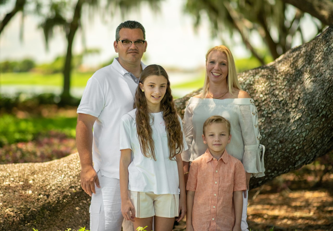 Family of four posing beside a large tree in a leafy park.