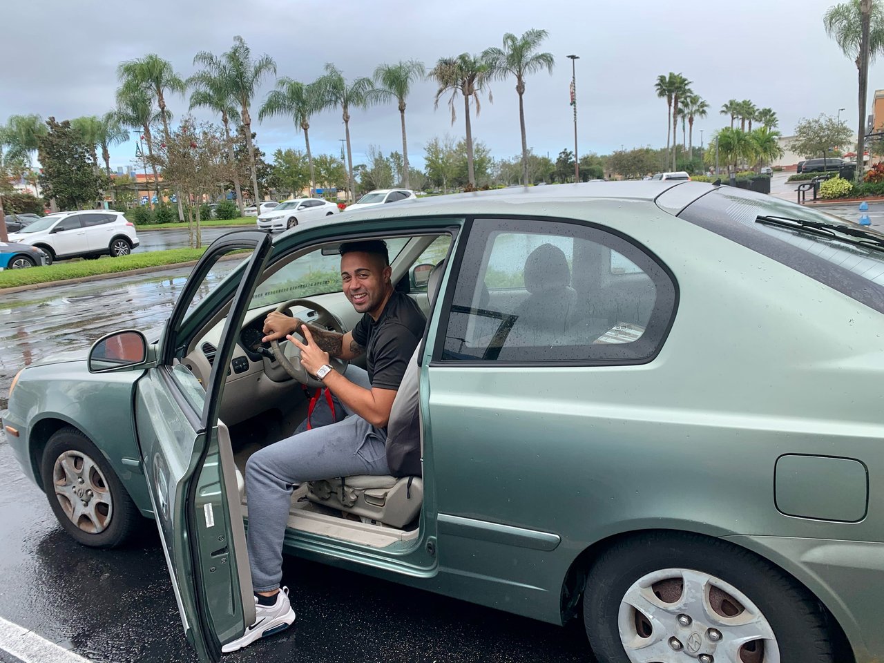 A smiling man flashes a peace sign from the open driver's seat of a green sedan.