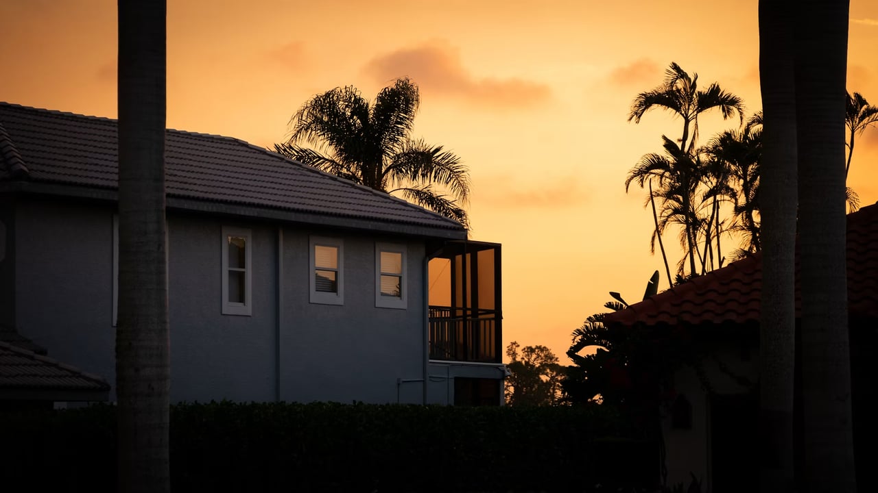 Two-story home with a balcony and palm trees silhouetted against an orange sunset.