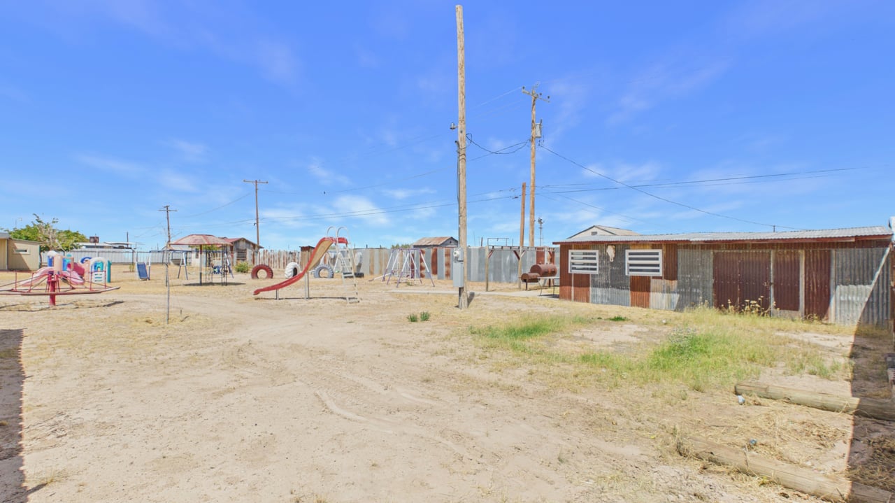 Sandy yard with playground equipment and metal-sided outbuildings.