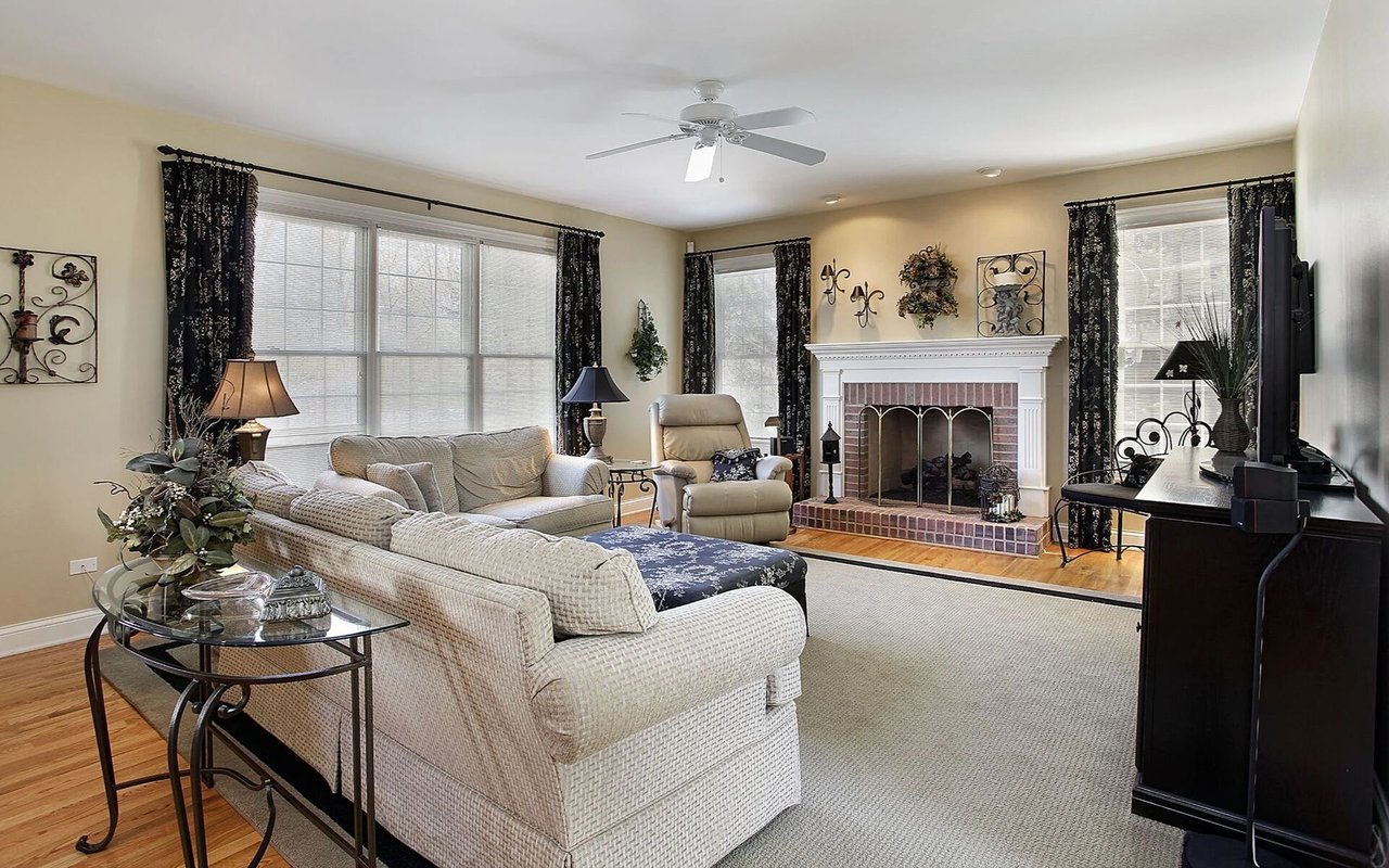 Living room with cream sofas, a brick fireplace, large curtained windows, and hardwood floors.