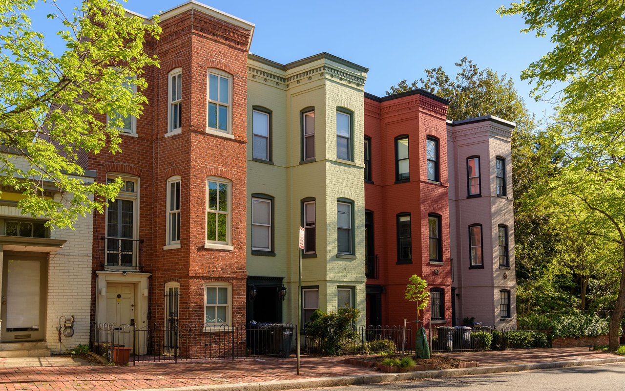 Colorful brick row houses with bay windows and leafy trees along a residential street.