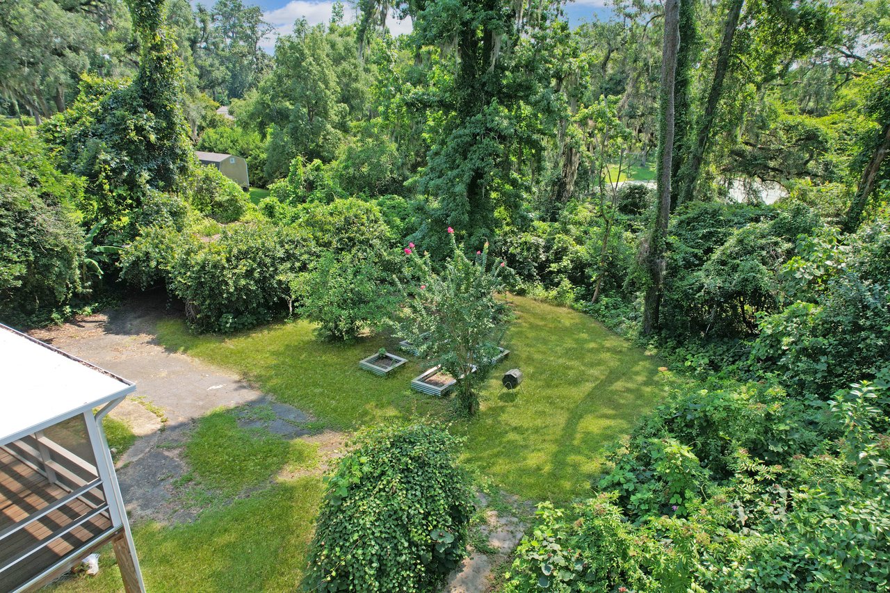 Lush green garden with tall trees and dense shrubbery surrounds a small grassy yard, featuring blooming shrubs and a canopied seating area on the lower left.