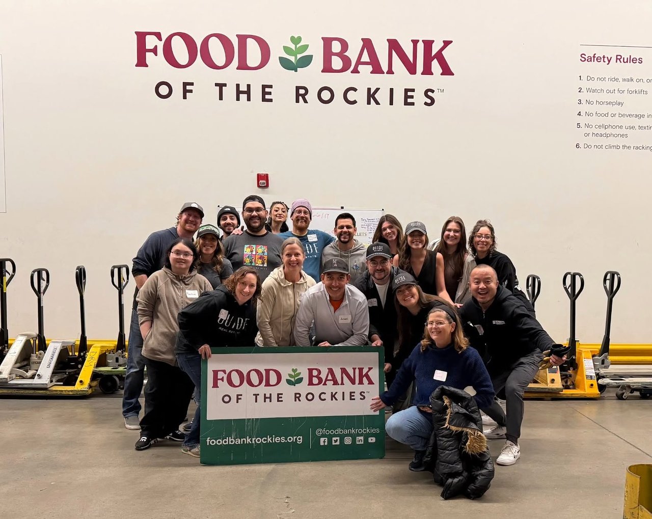 Guide Real Estate volunteers pose with a Food Bank of the Rockies sign in a warehouse.