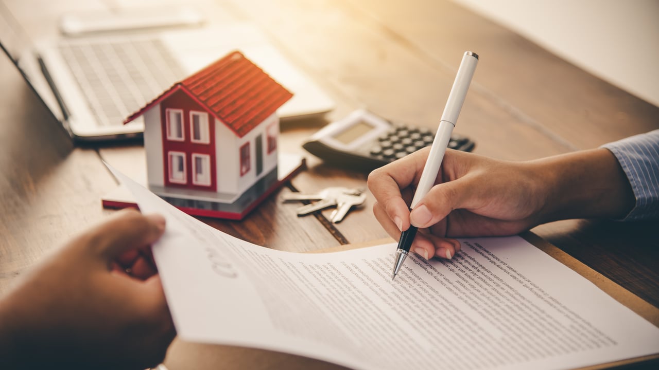 A person signs a home purchase contract beside a model house and keys.